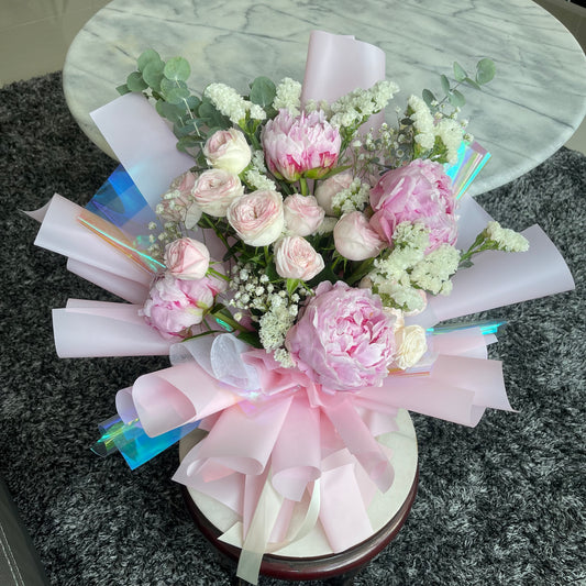 A floral arrangement featuring pink peonies and roses with light pink and iridescent wrapping.