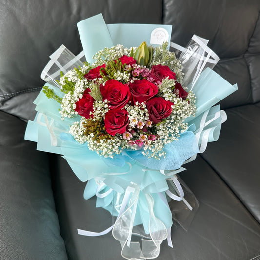 A bouquet of red roses wrapped in turquoise paper with baby's breath and accent flowers.