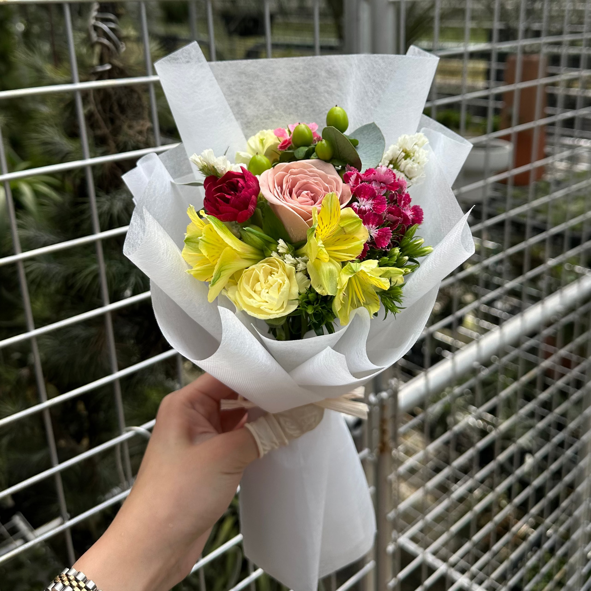Bouquet of nude toned rose, with mixed flowers held by a hand in front of a wire fence