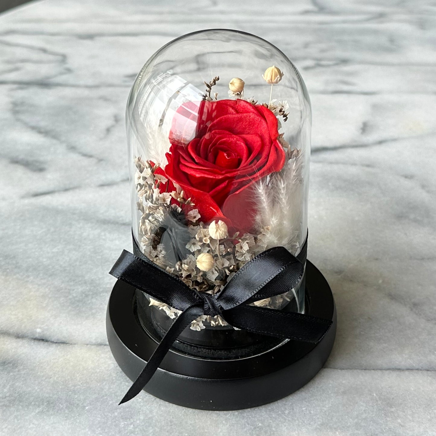 A single preserved red rose displayed inside a glass dome, placed on a black circular base with a black ribbon bow, set against a marble background.
