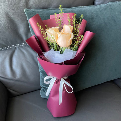 A bouquet of champagne roses wrapped in red paper with greenery, tied with a white ribbon.