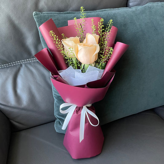 A bouquet of champagne roses wrapped in red paper with greenery, tied with a white ribbon.