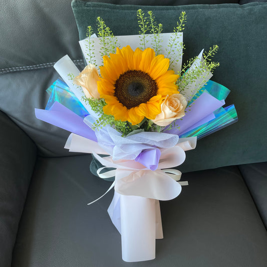 A bouquet of flowers including a sunflower and two champagne roses, wrapped with purple and white ribbons.