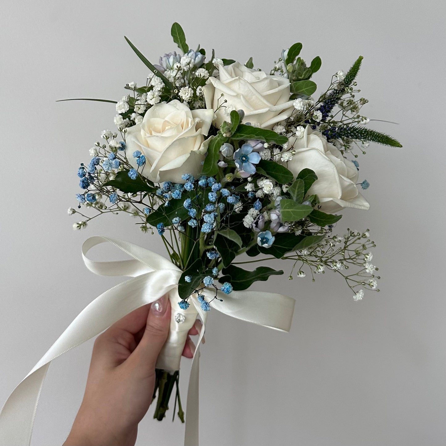A bridal Bouquet of white roses and greenery with a hand holding it against a plain background, with a long thick white ribbon.