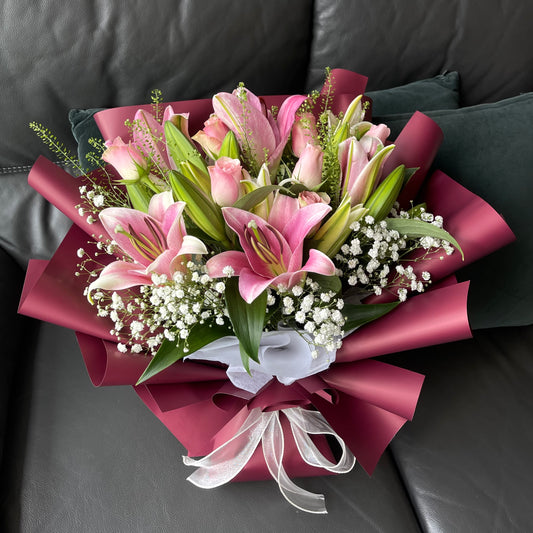 A bouquet of pink roses, lilies, and baby's breath wrapped in red paper with a white ribbon.