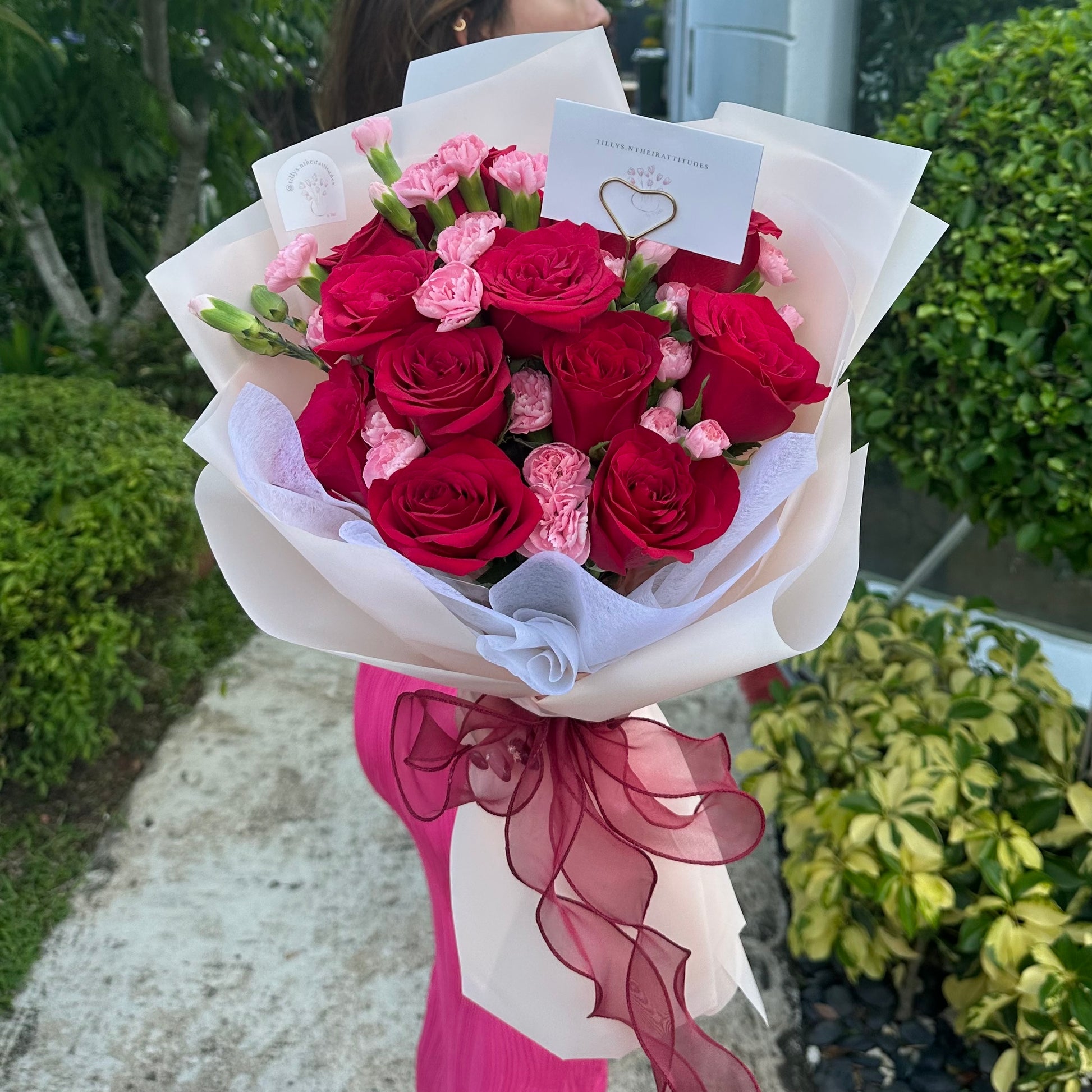 A dozen red roses paired with soft pink carnations in a bouquet, wrapped with white paper and a red ribbon, with a message card attached.