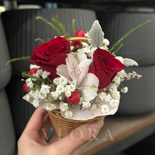 Hand holding a small bouquet of red roses and white flowers in a wicker basket with 'Viora' branding.