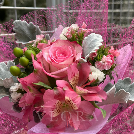Bouquet of pink flowers with greenery on a pink floral wrap, featuring the brand 'Vlora'.
