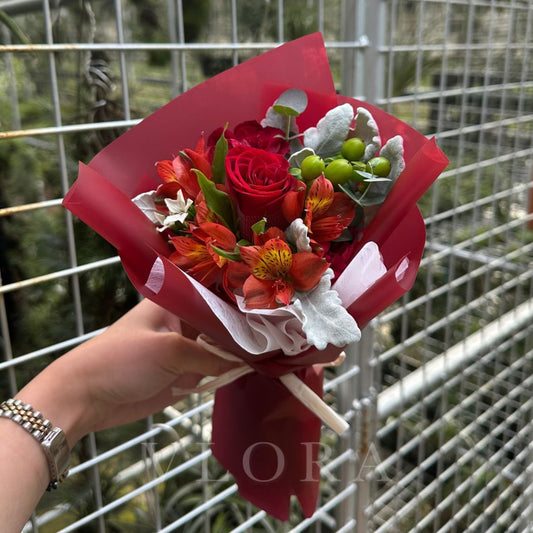 Bouquet of red and orange alstroemeria flowers with green leaves held by a person against a wire fence background.