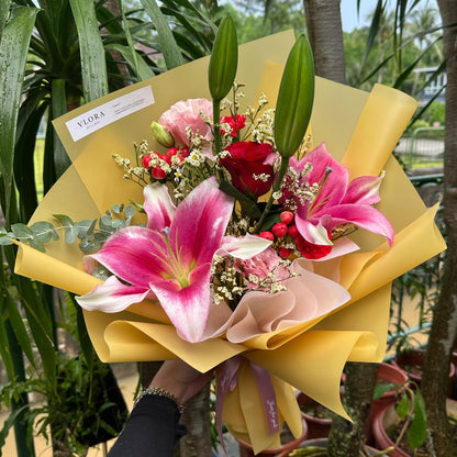 Bouquet of lily flowers with a yellow wrap held by a person, surrounded by potted plants.