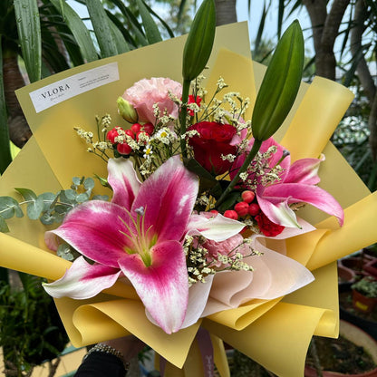 Bouquet of flowers with pink lilies and red flowers wrapped in yellow paper, set against a natural background.