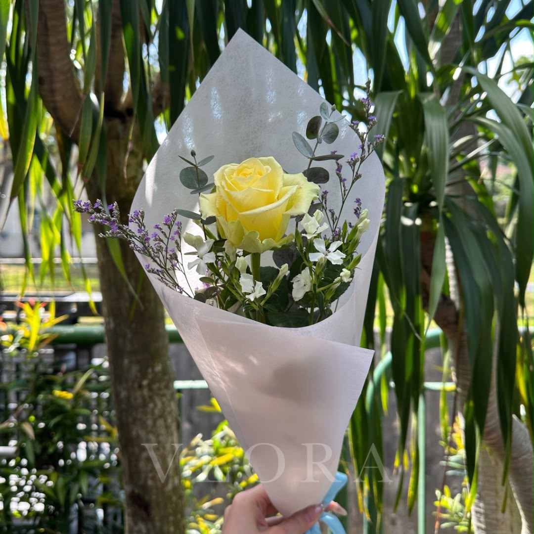 A single yellow rose bouquet wrapped in white tissue paper, held by a hand against a background of green palm trees.