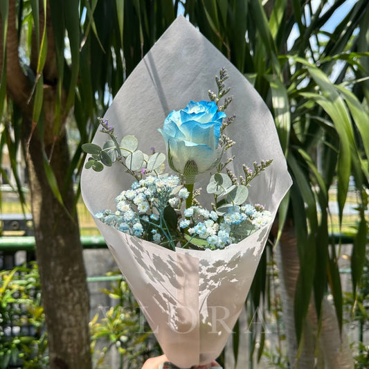A single wrapped bouquet featuring a blue rose, baby's breath, and eucalyptus leaves, wrapped in textured white paper and held outdoors in front of palm trees.