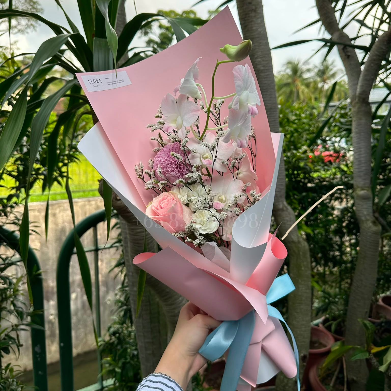 A hand holding a wrapped flower bouquet outdoors. The bouquet features white and pink flowers with green stems, wrapped in pink paper with a light blue ribbon bow.