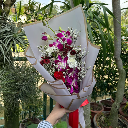 A hand holding a wrapped flower bouquet with purple orchids, white roses, and red accents, set against a lush green garden background.