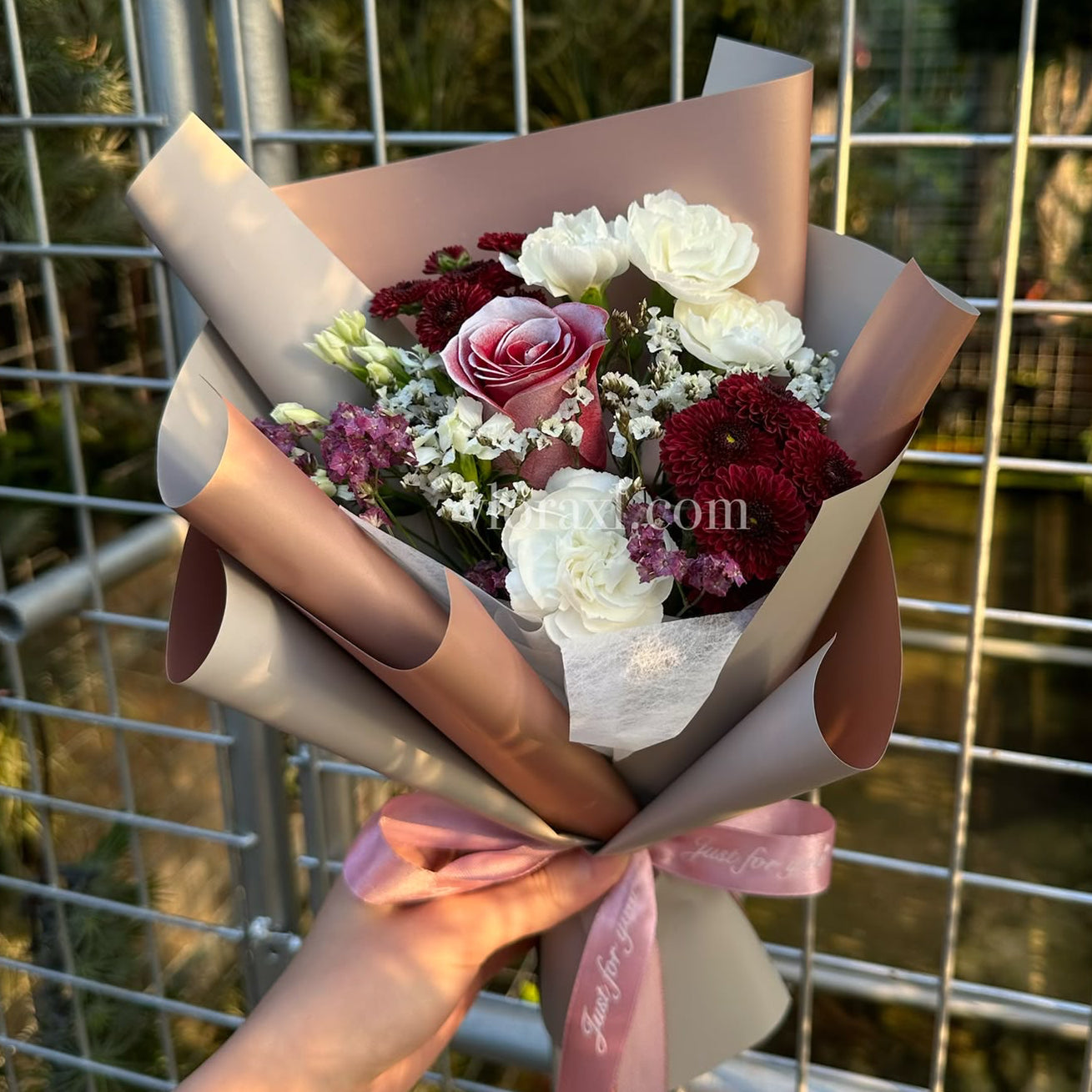 A hand holding a bouquet of flowers wrapped in blush pink paper, featuring white and burgundy roses with small white filler flowers, set against a metal grid fence background.