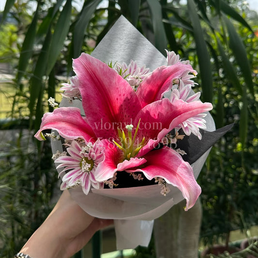 A hand holding a small bouquet of pink lilies and white daisy-like filler flowers wrapped in white paper, with a real garden background featuring green plants and trees.