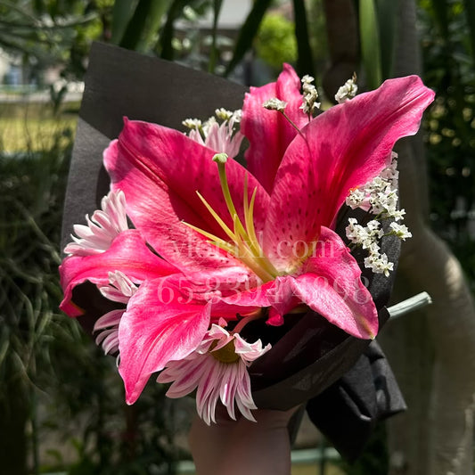 A hand holding a wrapped pink lily flower bouquet outdoors in a garden setting with green plants and trees in the background.