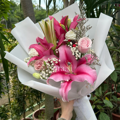 A hand holding a vibrant pink and white flower bouquet wrapped in white paper, featuring lilies, roses, and baby's breath, set against a background of potted plants.