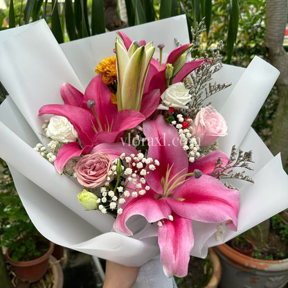 A vibrant flower bouquet held by a hand, featuring bright pink lilies, pink and white roses, white baby's breath, and a small yellow accent flower, wrapped in white paper against a background of potted plants.