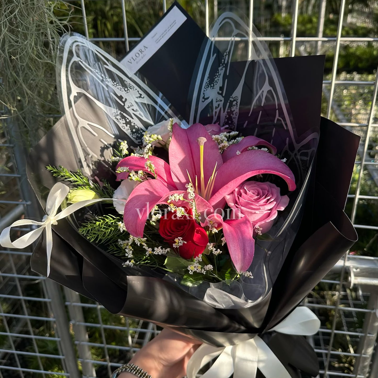 A hand holding a bouquet of pink lilies, pink roses, and red roses wrapped in black paper with white ribbon bows, in front of a metal grid fence.