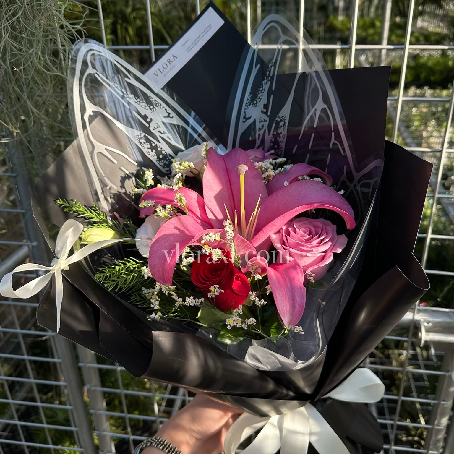 A hand holding a bouquet of pink lilies, pink roses, and red roses wrapped in black paper with white ribbon bows, in front of a metal grid fence.