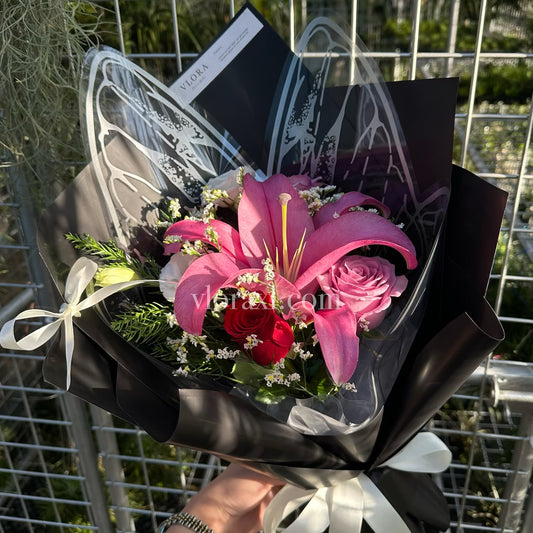 A hand holding a bouquet of pink lilies, pink roses, and red roses wrapped in black paper with white ribbon bows, in front of a metal grid fence.