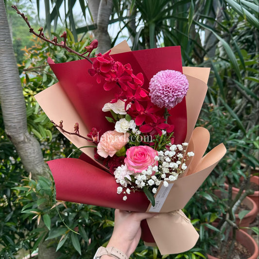 A hand holding a wrapped flower bouquet with red and pink flowers, white baby's breath, and red paper wrapping, set against a background of potted plants and trees.