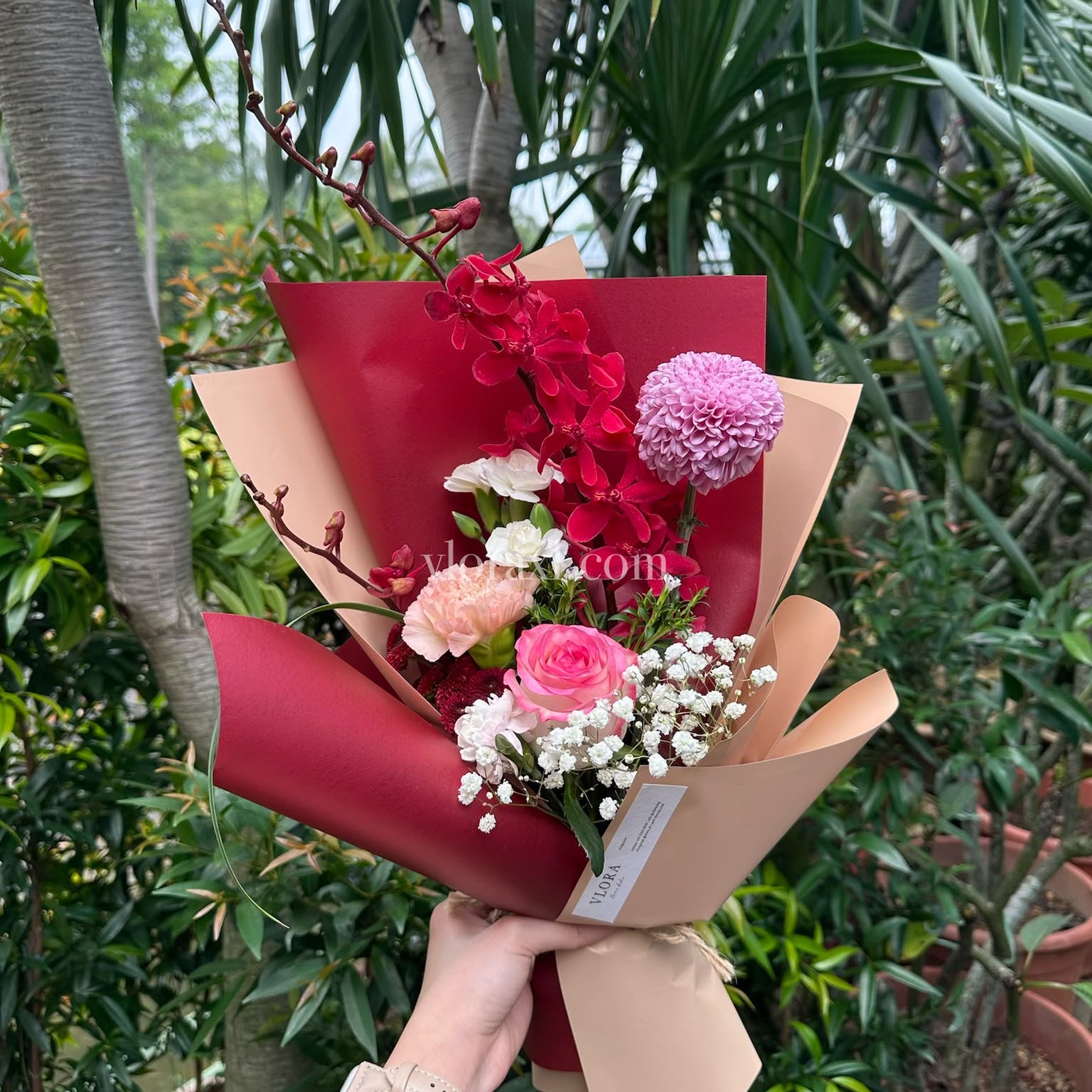 A hand holding a wrapped flower bouquet with pink roses, red orchids, white baby's breath, and a pink pom pom, set against a background of green potted plants.