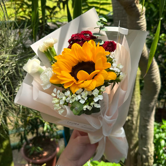 A hand holding a small floral bouquet wrapped in cream paper, featuring a large yellow sunflower, red carnations, white roses, and white baby's breath, set against a lush green garden background.