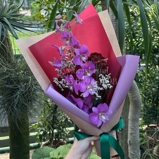 A hand holds a wrapped bouquet of purple orchids, white filler flowers, and a dark red rose in a garden setting with green plants in the background. The bouquet is wrapped in kraft paper with a bright red inner sheet, light purple paper, and a green ribbon.