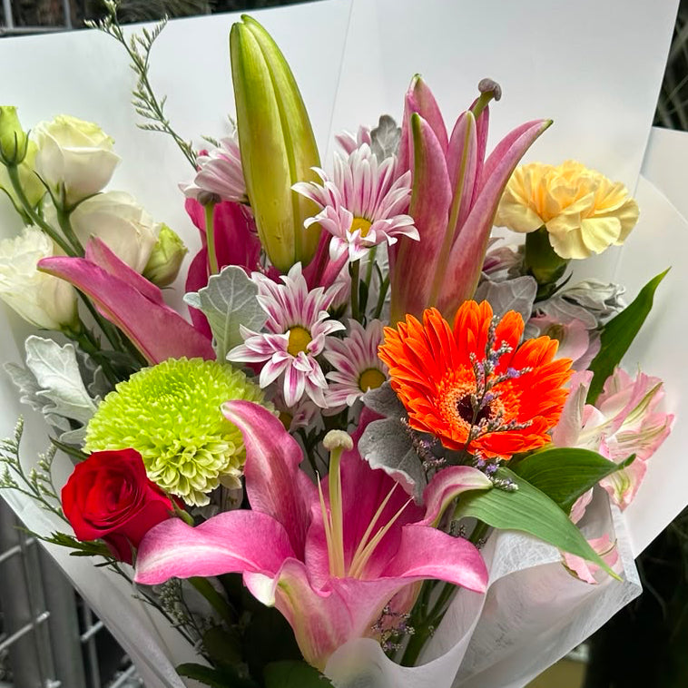 A vibrant mixed flower bouquet held by a hand, wrapped in white paper with a purple ribbon. The bouquet features pink lilies, an orange gerbera daisy, green hydrangea buds, and white roses, set against a background of a greenhouse or garden with metal shelving and potted plants.