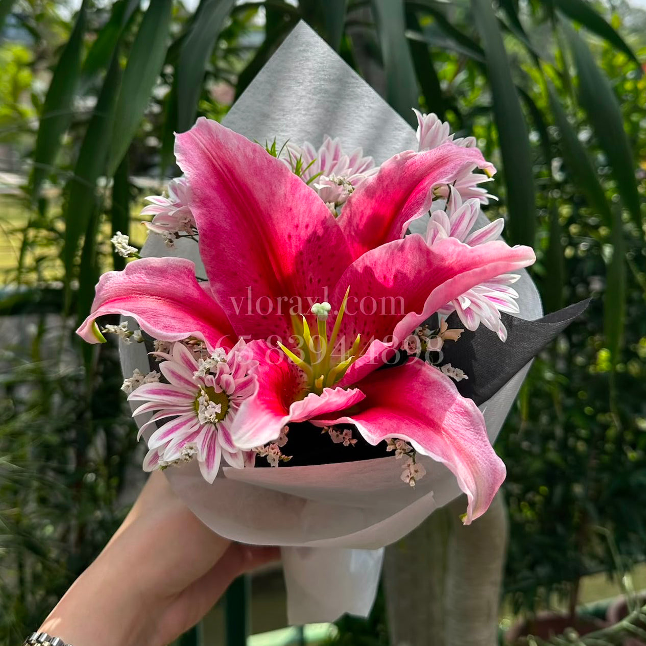 A hand holding a small bouquet of pink lilies and white daisy-like filler flowers wrapped in white paper, with a real garden background featuring green plants and trees.
