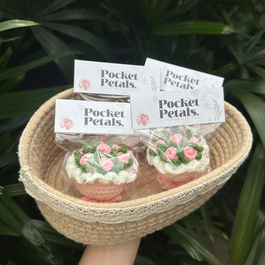 Two small crochet pink roses bouquet  labeled 'Pocket Petals' in a woven basket with green foliage in the background.