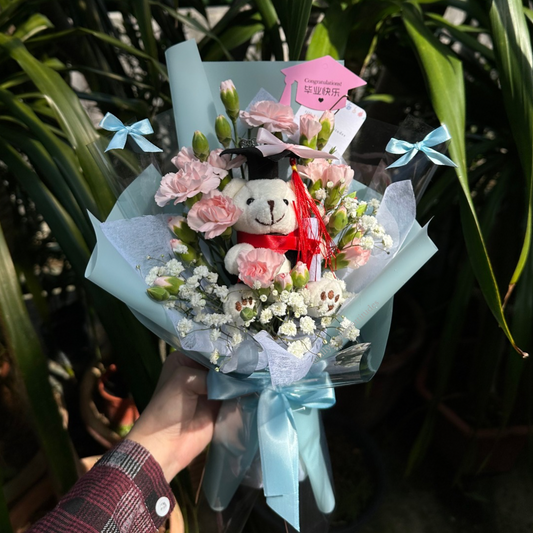 Bouquet with a teddy bear and pink carnation flowers held by a person, surrounded by greenery.