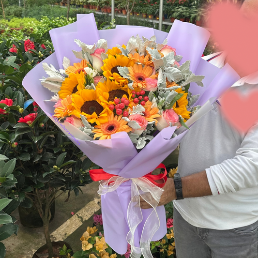 a large Bouquet of sunflowers and other flowers wrapped in purple paper held by a person outdoors.