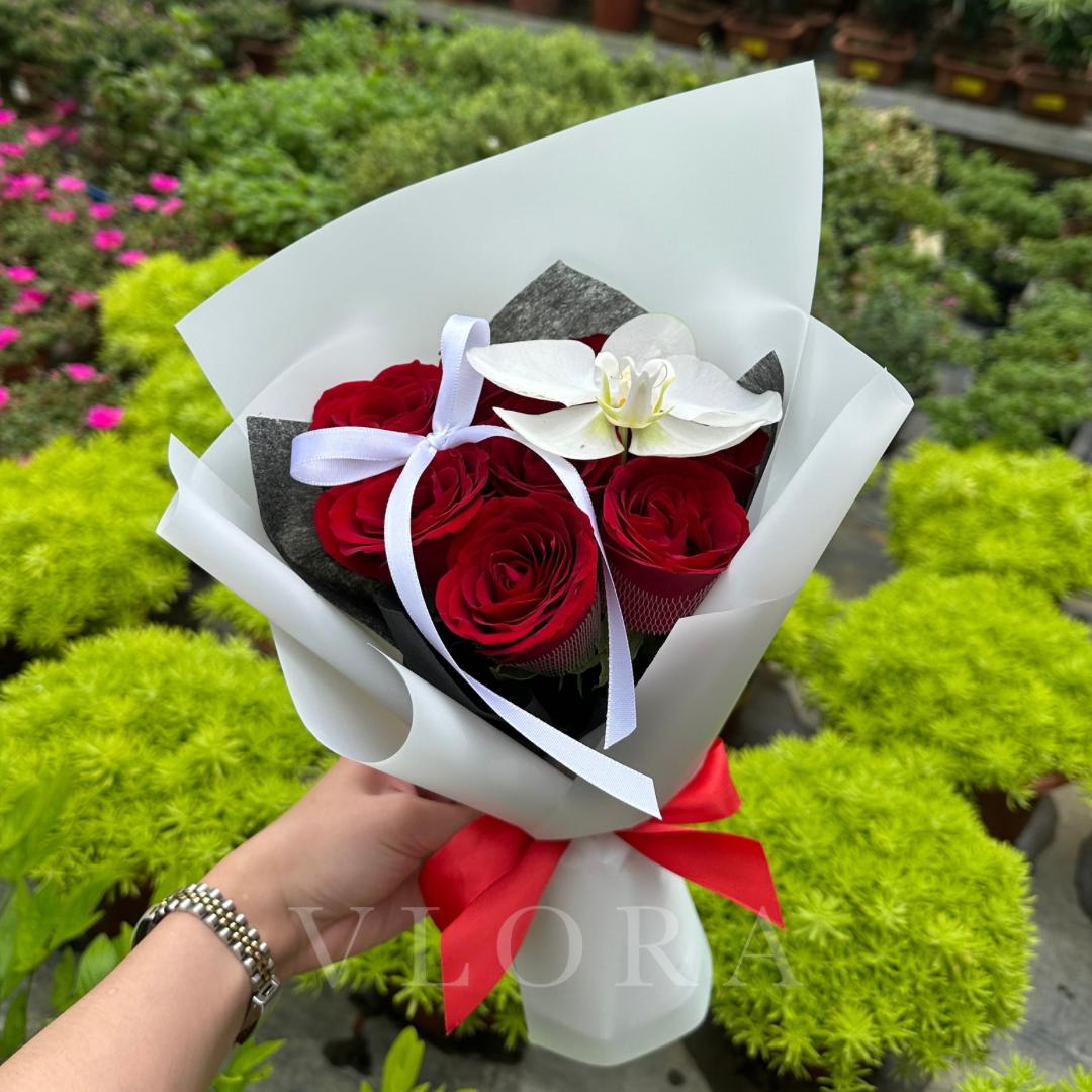 Bouquet of red roses and a white orchid wrapped in white paper with a red ribbon, held by a person in a garden setting.