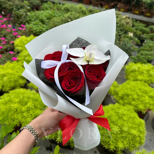 Bouquet of red roses and a white orchid wrapped in white paper with a red ribbon, held by a person in a garden setting.