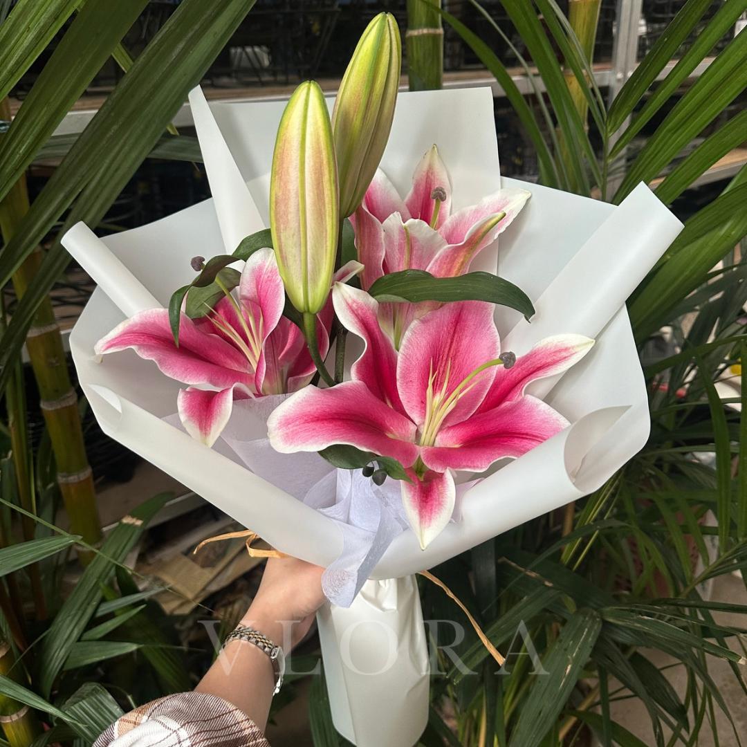 Bouquet of pink lilies in a white paper wrap held by a person with green plants in the background.