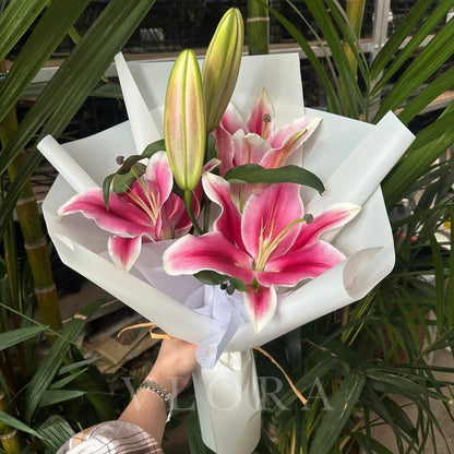 Bouquet of pink lilies in a white paper wrap held by a person with green plants in the background.