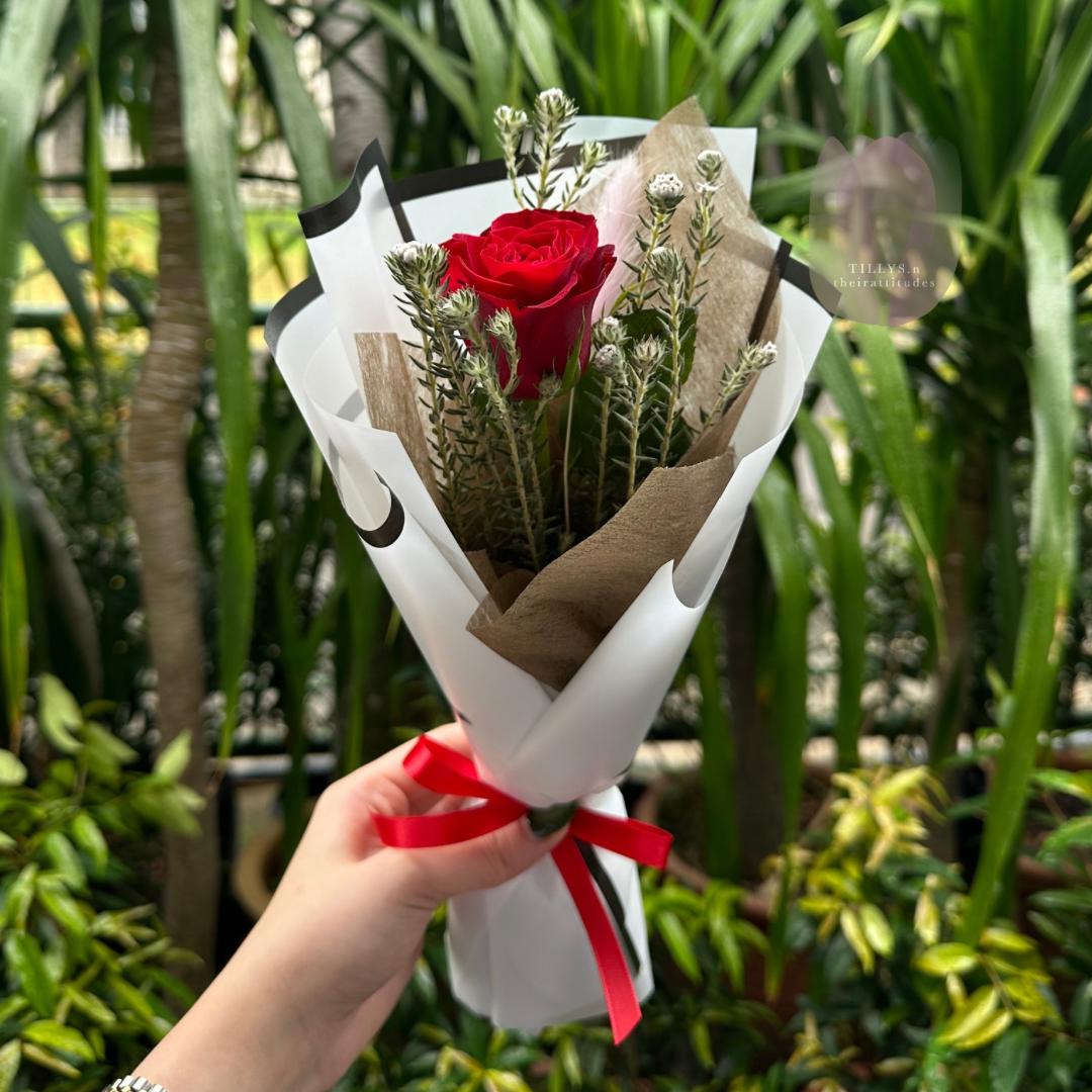 Bouquet of red roses with greenery wrapped in brown paper and white paper, held by a hand against a green foliage background.