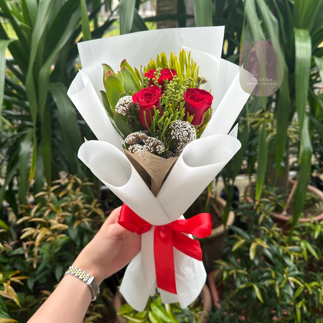 Bouquet of red roses and greenery wrapped in white paper with a red ribbon, held against a green leafy background.