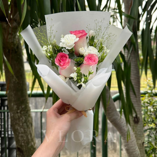 Bouquet of pink and white flowers wrapped in white paper held by a hand with a blurred green background.