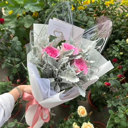 Bouquet of pink roses and silver foliage with a clear plastic wrap, held by a person among potted plants.
