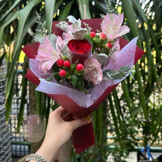 Bouquet of flowers with red and pink tones held by a hand against a green leafy background.