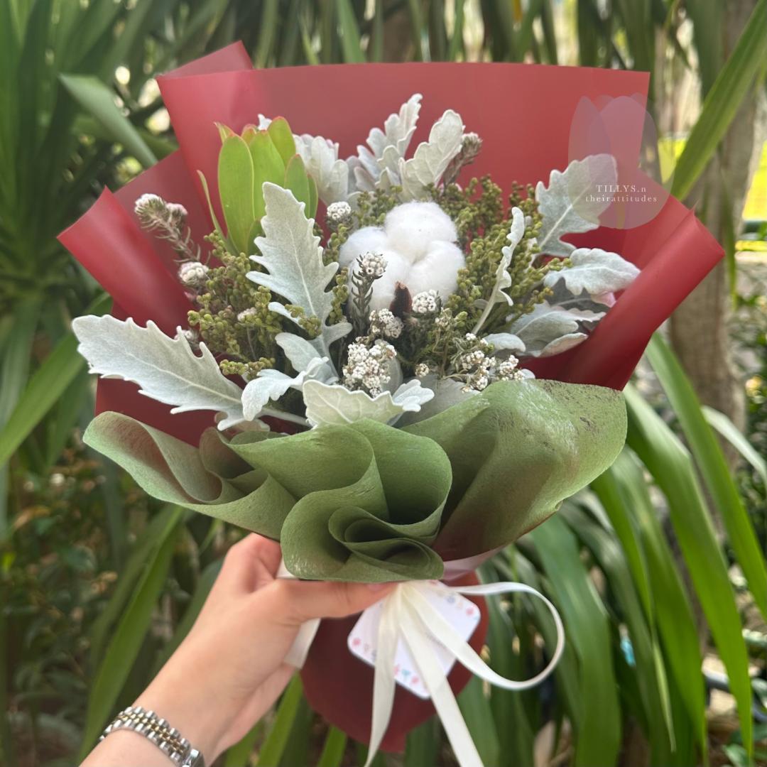 Bouquet of flowers with greenery and white elements held by a person against a natural background.
