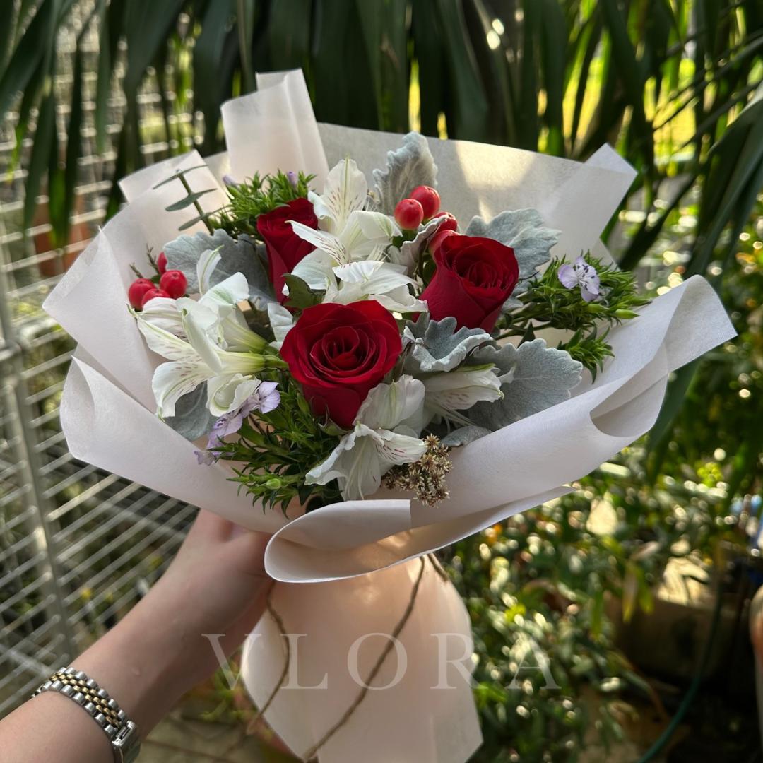 Bouquet of red roses and white flowers wrapped in white paper, held by a person with a blurred background.