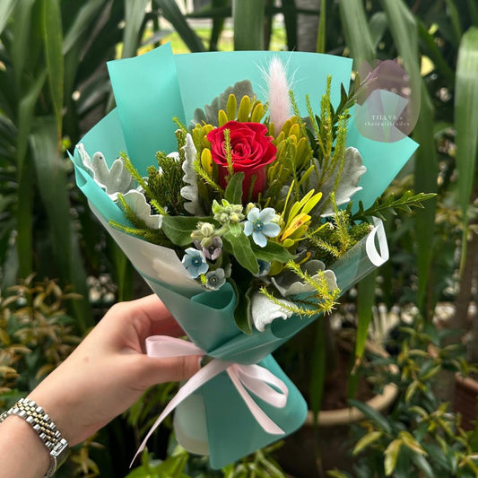 Bouquet of flowers wrapped in turquoise paper with a pink ribbon, held by a hand against a green foliage background.