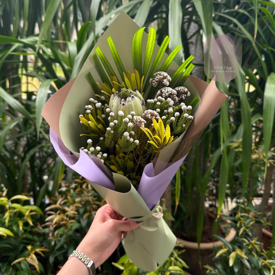 Bouquet of flowers held by a hand with a green and purple wrap against a plant background