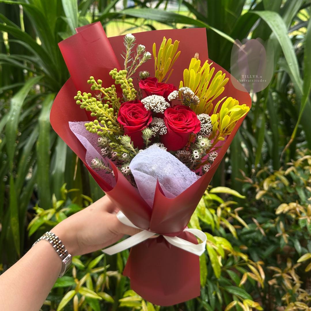 Bouquet of red and yellow flowers wrapped in red paper held by a person against a green leafy background.